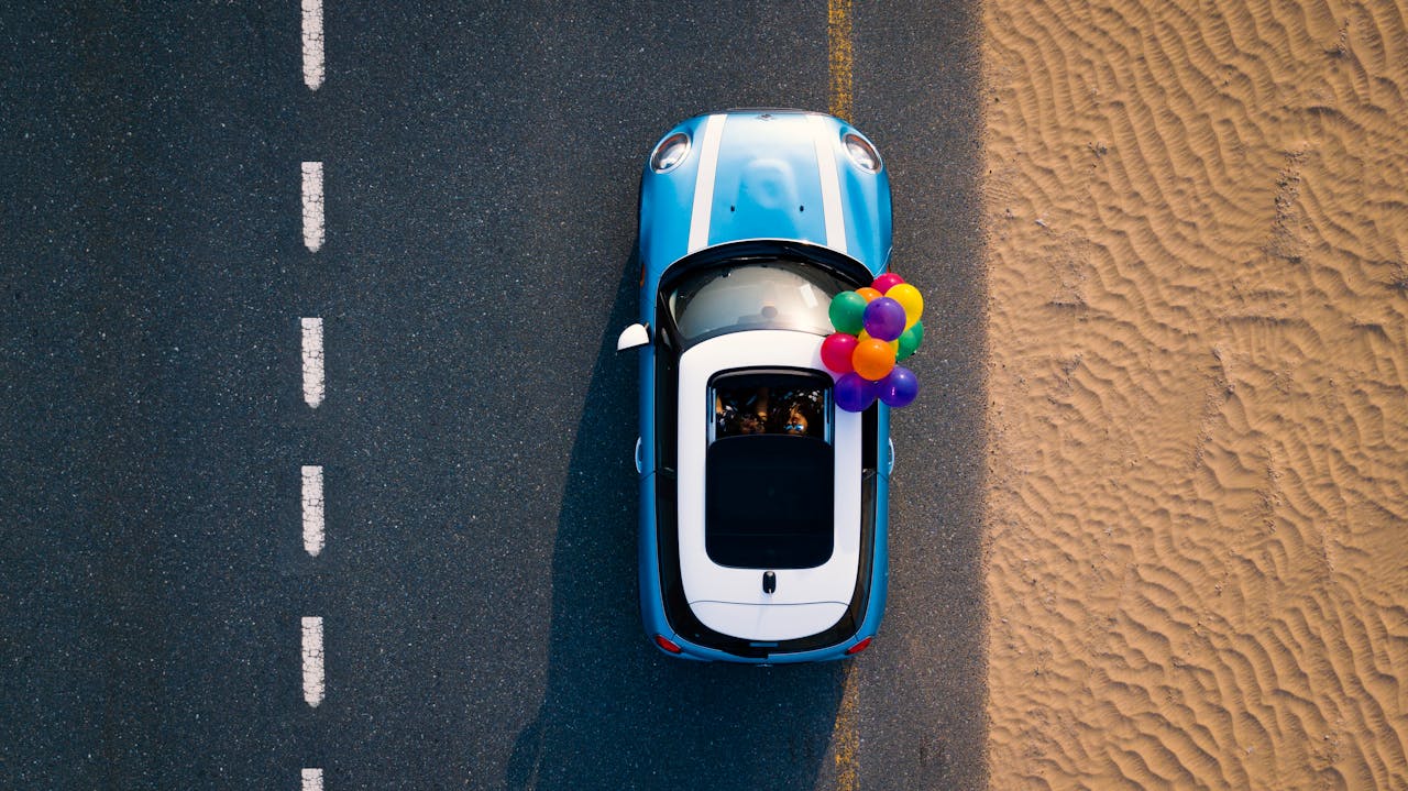 ours-journey A Mini Cooper with colorful balloons drives along a desert road. Captured from above.