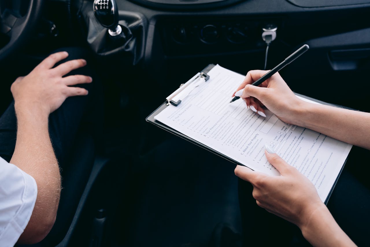 about-img Close-up of a person writing on a clipboard inside a car, showing hands and a gear shift.