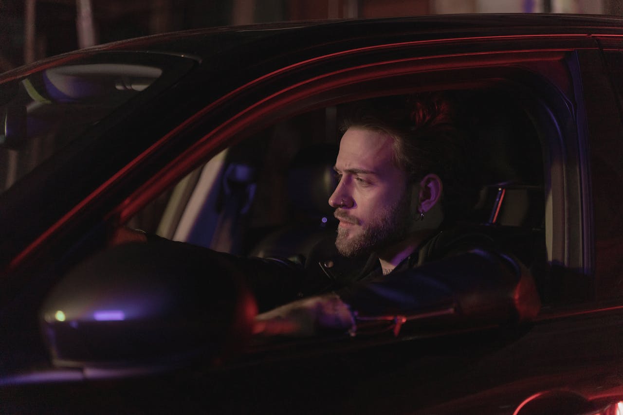 services-03 Bearded man drives a car at night, illuminated by red neon lights, creating a moody atmosphere.